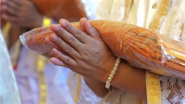 Participants in white clothing line up holding folded orange robes as soft sunlight reveals intricate lace details and hands within a medium to large ceremonial setting.