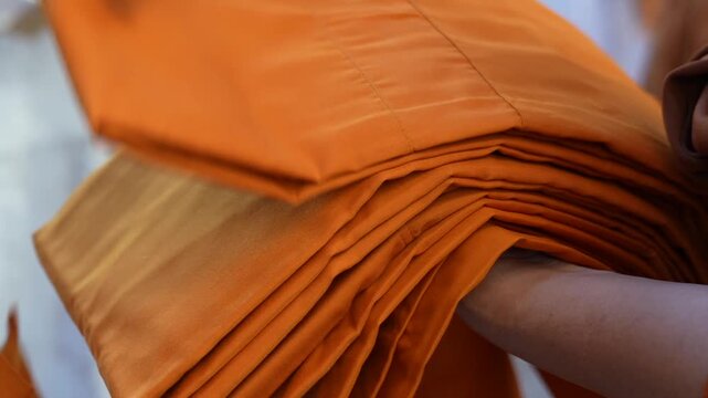 A senior monk adjusts a novice robe as hands tighten the woven belt around orange cloth in sunlight with focus on texture folds and precise fitting.