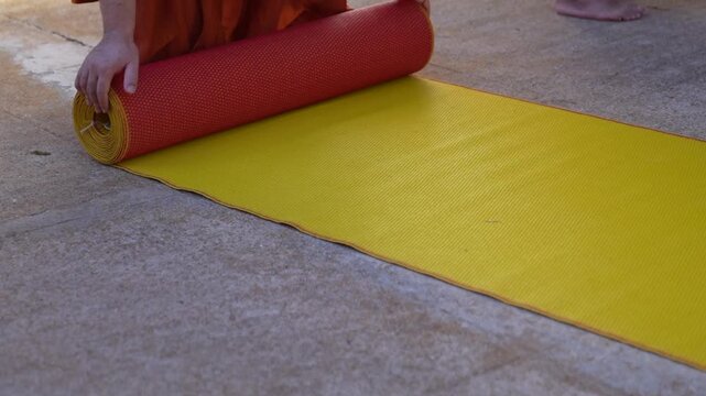 A monk rolls up a yellow mat on the concrete ground. Orange robe fabric and hands frame the storage action.
