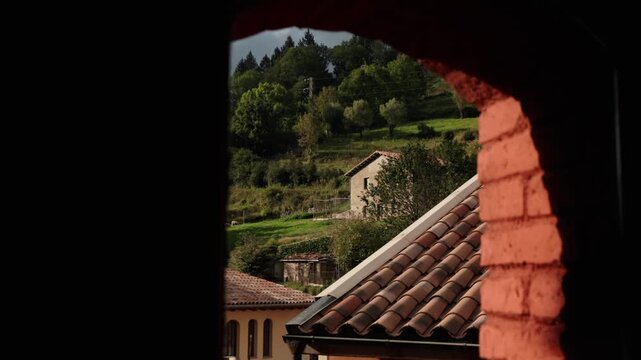 Static shot from inside a house window showing tiled roof, village homes and green hillside in Camprodon Spain