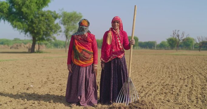 Indian mature old aged two lady woman standing look camera outside day rural area. Happy worker female hold farm rake in hand staring cam do pose enjoy desi life village ground place 