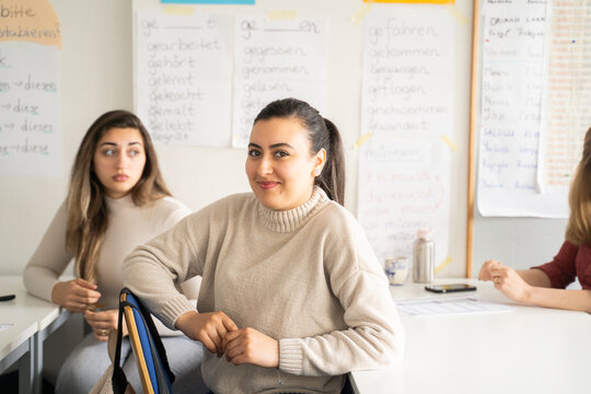 Two female students with long hair sitting at desks in a multicultural classroom setting