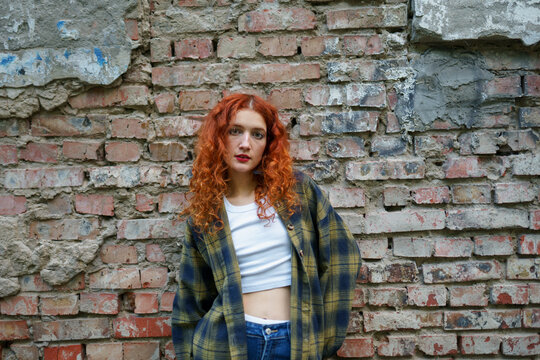 Red haired woman in white top and jeans posing by old brick wall