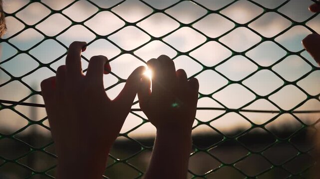 Hands gripping chainlink fence at sunset with fingers through mesh. Person holds fence barrier outdoors. Silhouette hands on chainlink at sunset. Fingers grip wire fence with sunlight behind.