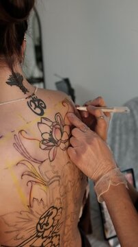 Close-up of a female tattoo artist draws a scketch of intricate floral and mandala design on a woman's upper back. Free hand tattoo technique.