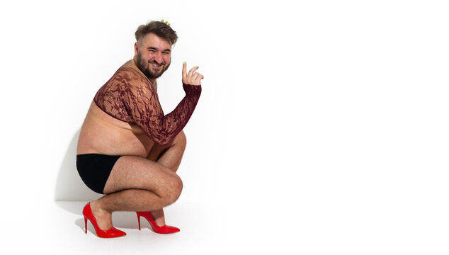Photo banner. Mature man with beard, big belly, black briefs, mesh through tank top, bare hairy legs, red stiletto high heels sits, squats on floor looks on camera on white. Pride month. Copy space