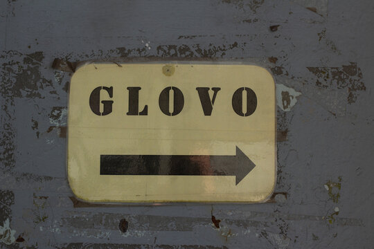 Tbilisi, Georgia - April 10, 2026: Horizontal street city photo. Sign board with Glovo logo and black arrow on gray metal wall. Concept of food, drink, lunch, dinner, job, pick-up point, job office