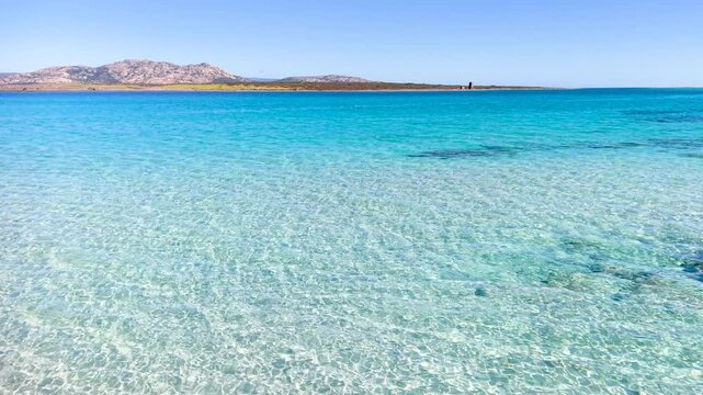 Cinematic low altitude drone flight skimming the surface of transparent turquoise water at La Pelosa beach in Sardinia showing white sand patterns and sunlight reflections on the sea