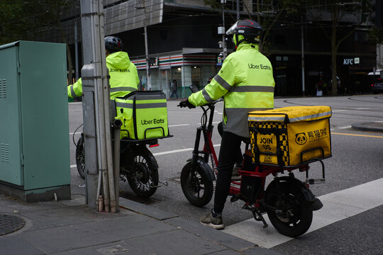 Pair of food delivery couriers on bikes, both wearing Uber Eats branded gear, sitting at an intersection in the city of Melbourne