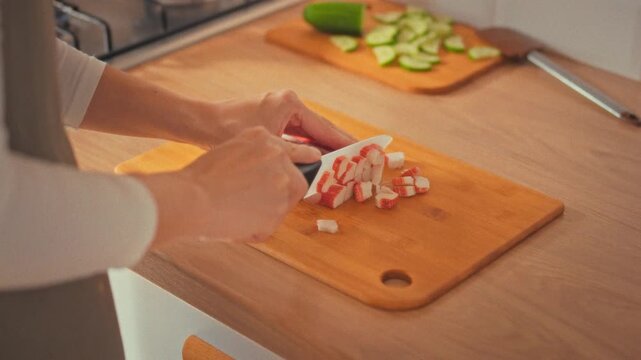 A woman, like a chef, deftly slices bright crab sticks on a kitchen counter, mixing them with fresh vegetables and herbs. The careful cooking process showcases home kitchen food preparation