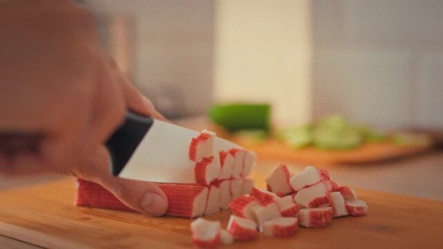 Cutting bright crab sticks, a woman chef slices the seafood with precision in a home kitchen, surrounded by fresh vegetables and spices, illustrating a cooking routine where skill meets meal prep