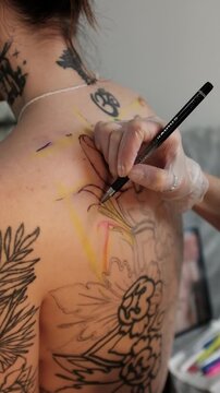 Close-up of a female tattoo artist draws a scketch of intricate floral and mandala design on a woman's upper back. Free hand tattoo technique.