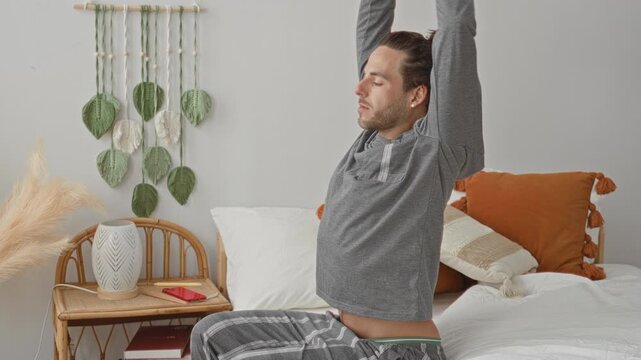 Man sitting on bed in gray loungewear reaching with right hand for red smartphone on wicker nightstand beside pillows and macrame wall hanging in studio; calm morning.