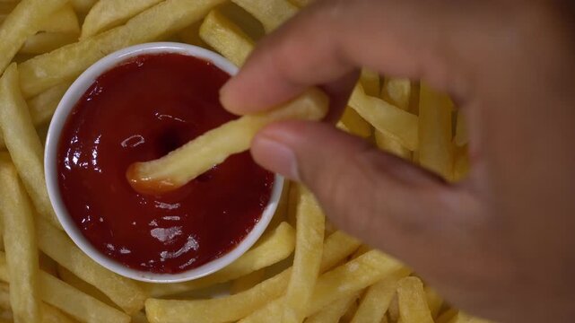 Top down view of a hand dipping a french fry into a ketchup bowl surrounded by fried potatoes