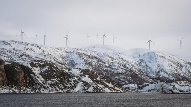 View of wind turbines standing tall on snow-dusted hills under a soft, overcast sky, creating a serene yet powerful landscape, Brensholmen, Troms, Norway.