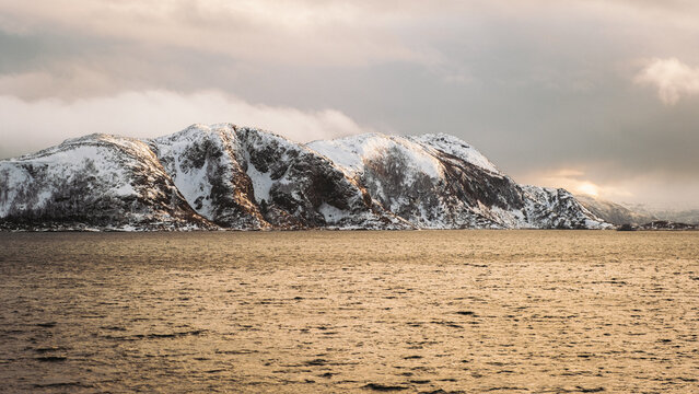 View of the sun reflecting off the water in front of snow-capped mountains under a cloudy sky, Brensholmen, Troms, Norway.