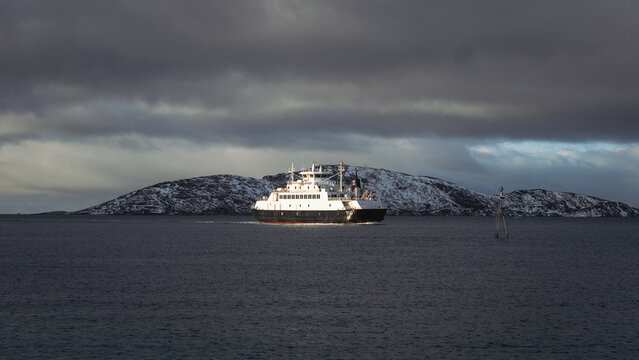 View of a ferry cutting through the still, dark waters toward a snow-dusted island under a moody, overcast sky, Brensholmen, Troms, Norway.