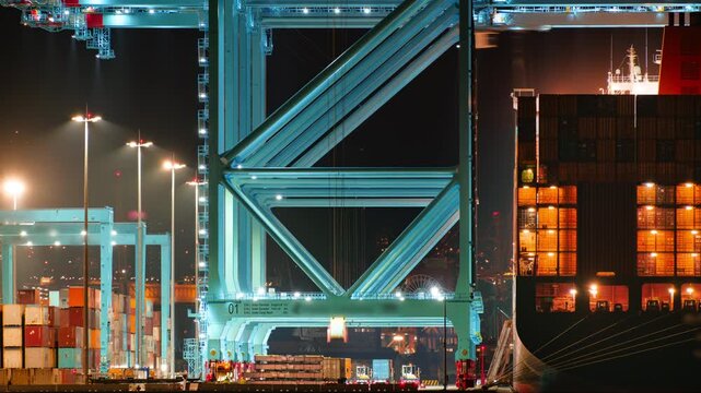 Timelapse of quay cranes lift and move shipping containers onto a large container ship during a nighttime operation at bustling cargo port. Semi-trucks moving in motion blur and gantry cranes working