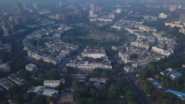Elevated aerial perspective reveals Connaught Place's distinctive circular Georgian-style architecture surrounding a central park in New Delhi, India, with urban sprawl and residential buildings.