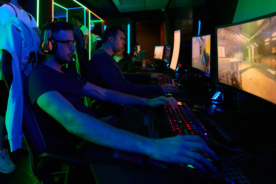 Competitive Gamers Playing Multiplayer Shooter on PCs in Neon-Lit Gaming Arena With Headsets and Keyboards