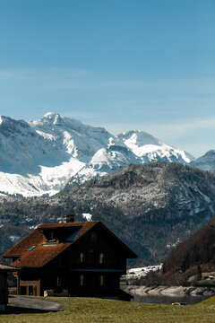 View of a charming Swiss chalet nestled against a backdrop of snow-capped mountains under a serene blue sky, captured from a moving train, Train from Lucerne to Interlaken, Switzerland.