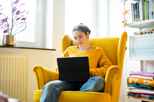 Woman relaxing in a yellow armchair at home with a laptop and a cup of tea