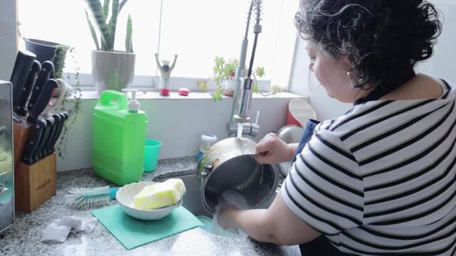 Mature Hispanic woman washing a cooking pot in the kitchen sink
