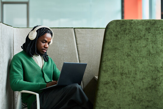 African American young man wearing headphones, working on laptop in office booth, focusing on remote tasks. Ideal for hybrid work, productivity, concentration, digital workflow marketing