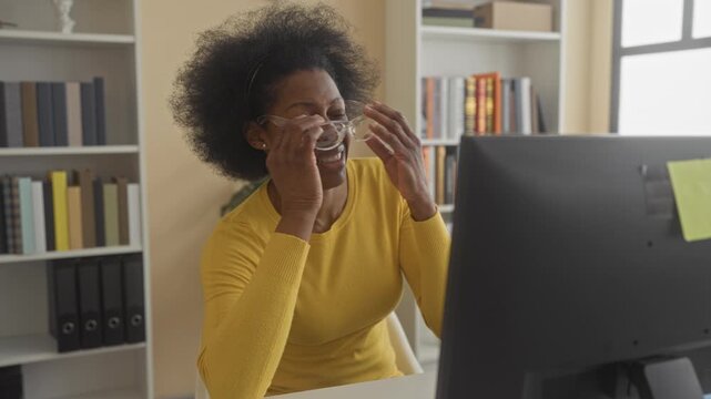 Woman wearing yellow sweater with hand to temple at computer monitor in building, bookshelf visible; stress concentration.