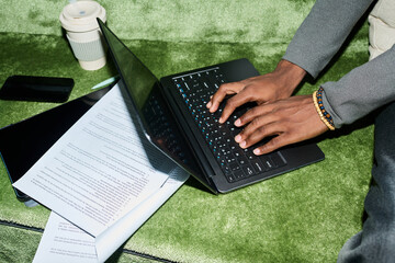 Naklejka na ściany i meble High angle view of Black young professional typing on laptop beside documents and smartphone on grass, working outdoors. Useful for remote work, productivity, freelance business marketing