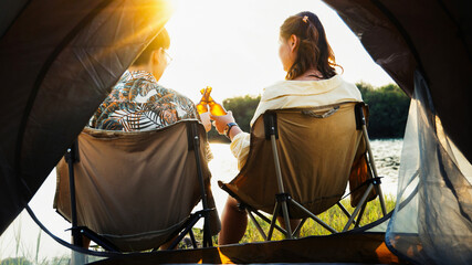 Couple seated comfortably in camping chairs sharing refreshing beverages while admiring peaceful lake scenery illuminated by bright evening sunlight outdoors © pixfly