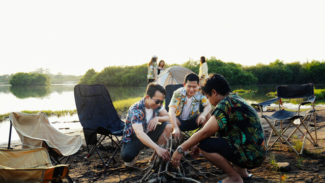 Friends building a wooden campfire together near their lakeside tent, enjoying warm sunlight and relaxed summertime camping moments outdoors.