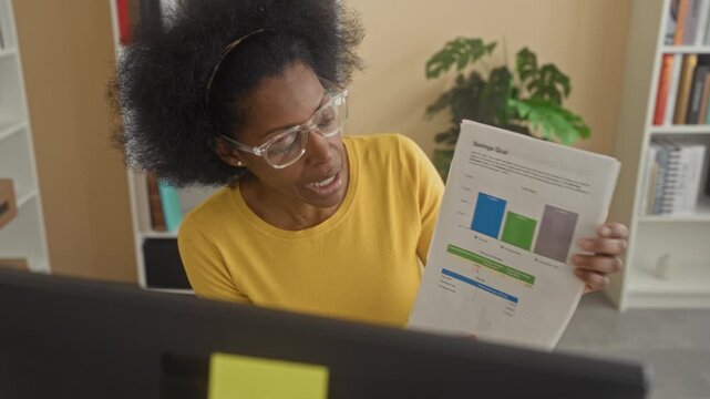 Woman in yellow sweater holds a company performance report and points pen to chart on computer monitor in building; analysis focus.