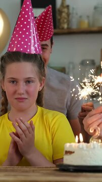 A girl in a yellow shirt and a pink party hat looks uncomfortable during a birthday celebration. Sparklers light up the scene as a cake is presented in the background.
