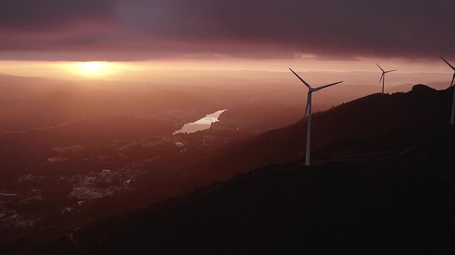 Three blade up wind turbines on Serra da Boneca in Penafiel Porto District Norte Portugal above Douro Valley with river reflecting sunset light across layered terrain, establishing aerial shot