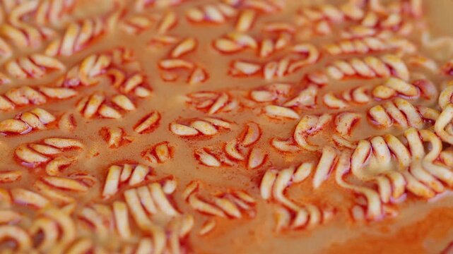 Closeup spicy instant noodles simmering in broth, bubbling orange broth with chili specks, curly strands softening, rising steam, homey warm atmosphere, quick comfort meal