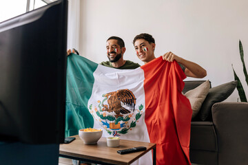 Obraz premium Mexican father and teenage son celebrating a football moment together at home. Happy family supporters cheering on the couch with national flag, snacks and drinks.