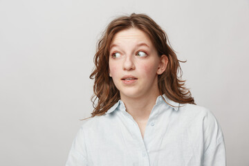 Obraz premium Young woman with curly hair wearing white shirt looks sideways with curious expression on light background. Concept of surprise, doubt, and casual lifestyle portrait.