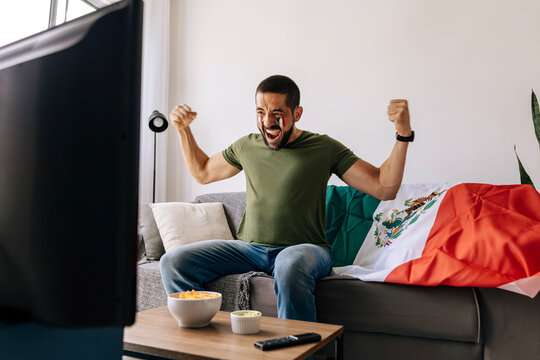 Mexican football supporter celebrating excitedly at home while watching a match on TV. Adult man cheering on the sofa with national flag, snacks and drinks.