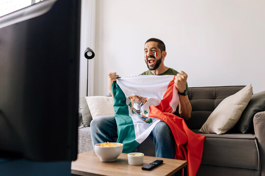 Mexican football fan celebrating at home while holding national flag and watching a match on TV. Adult man cheering on the sofa with snacks and drinks in the living room.