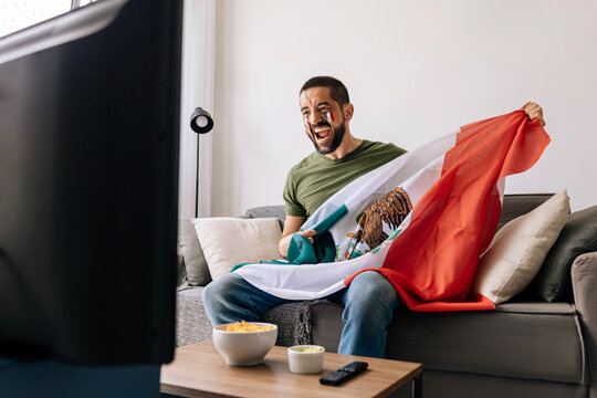Mexican football fan celebrating at home while holding national flag and watching a match on TV. Adult man cheering on the sofa with snacks and drinks in the living room.