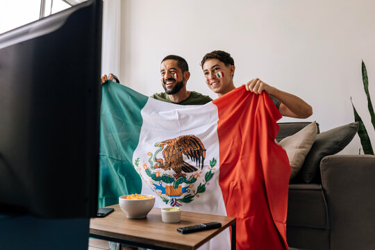 Mexican father and teenage son celebrating a football moment together at home. Happy family supporters cheering on the couch with national flag, snacks and drinks.