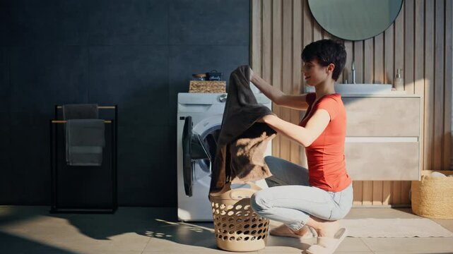 Young woman separating garments putting laundry in machine drum weekend morning