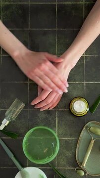 Top view of woman's hands over a variety of skincare products, dipping finger into cream jar on dark tile background