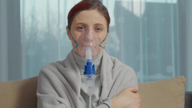 A woman sits in a room wearing a mask connected to a nebulizer. She inhales steam from a saline solution to help with respiratory issues like coughs and allergies.