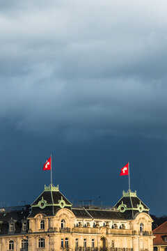Zurich, Switzerland - 02 April 2023: View of Swiss flags atop a grand building, contrasting sharply against the ominous, dark sky above.