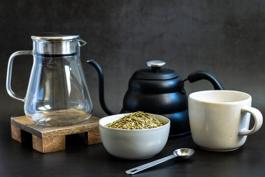 Yerba Mate loose tea in a white ceramic bowl with kettle and tea pitcher on a dark backdrop