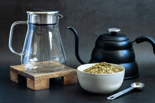 Yerba Mate loose tea in a white ceramic bowl with kettle and tea pitcher on a dark backdrop