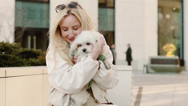 4K slow motion stylish woman sitting outdoors in city holding cute Maltese dog, urban lifestyle, pet companionship, modern architecture background, calm emotional scene