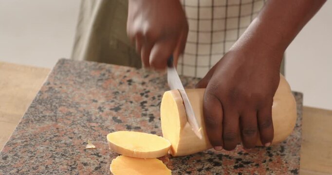 African American man at granite counter wearing apron slicing butternut squash with blue chef knife